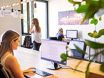 Bürosituation, Frau und Mann sitzen am Computer während eine andere Frau telefoniert.