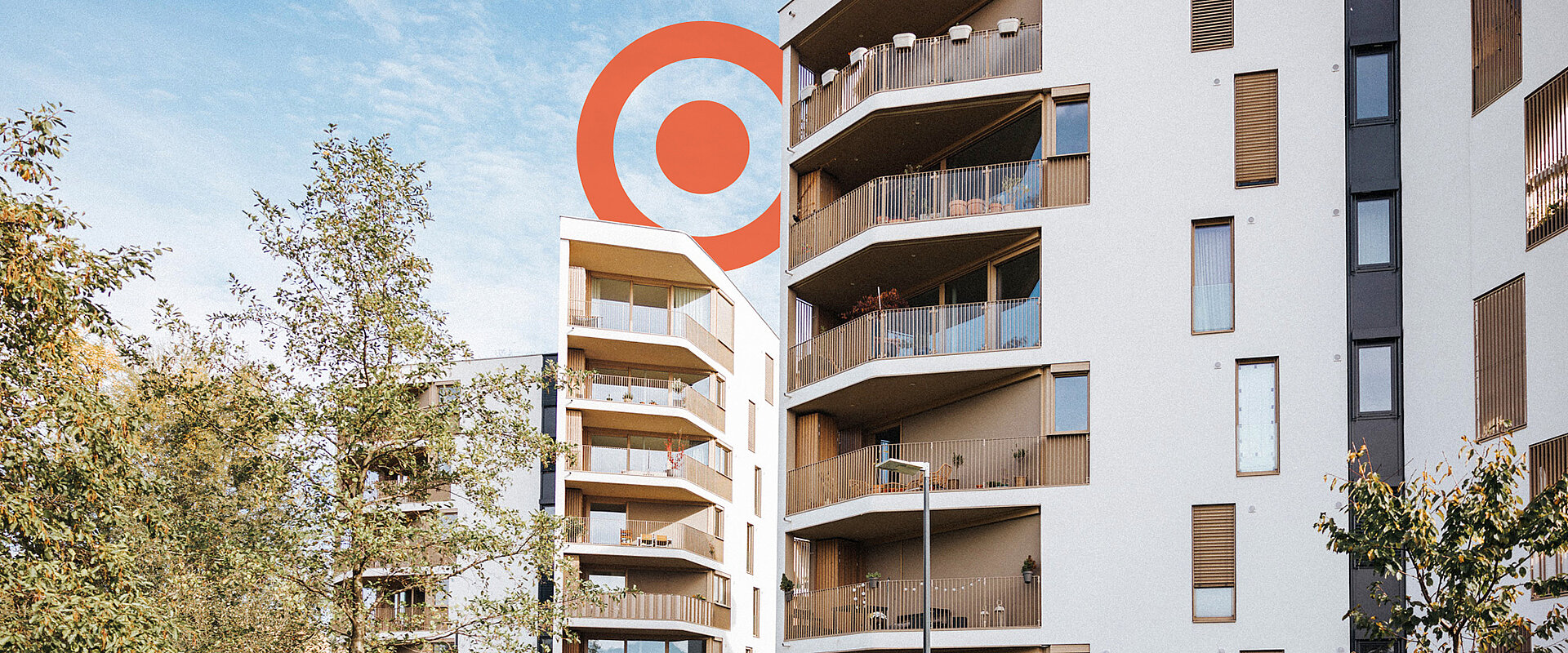 Foto von den Neubauten des Quartier Rauchmühle in Salzburg mit orangem PRISMA Logoelement im Hintergrund.